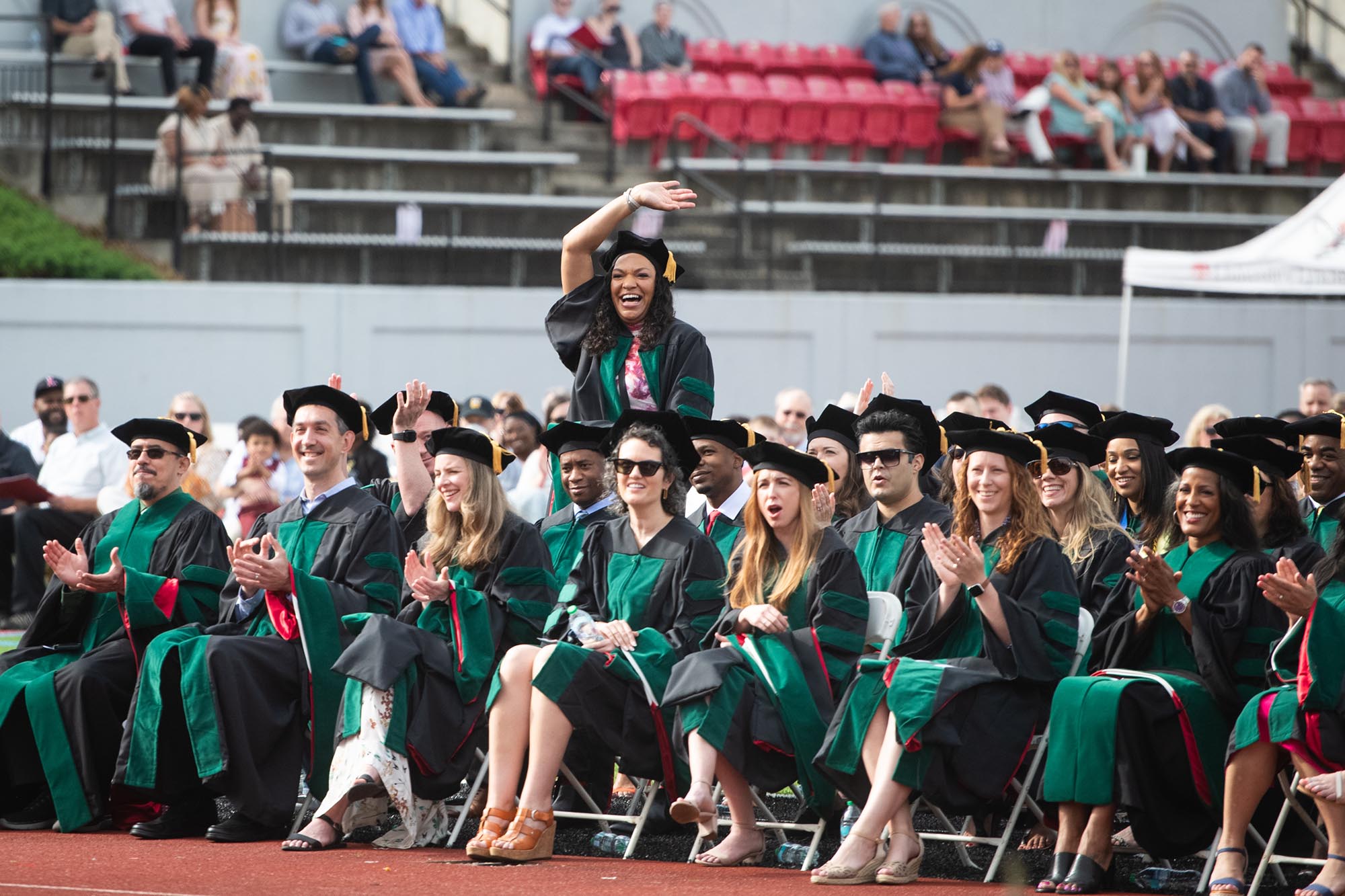 Woman in doctorate regalia standing among other graduates and waving.