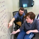 A professor and a student in casual clothing sit and write on a whiteboard