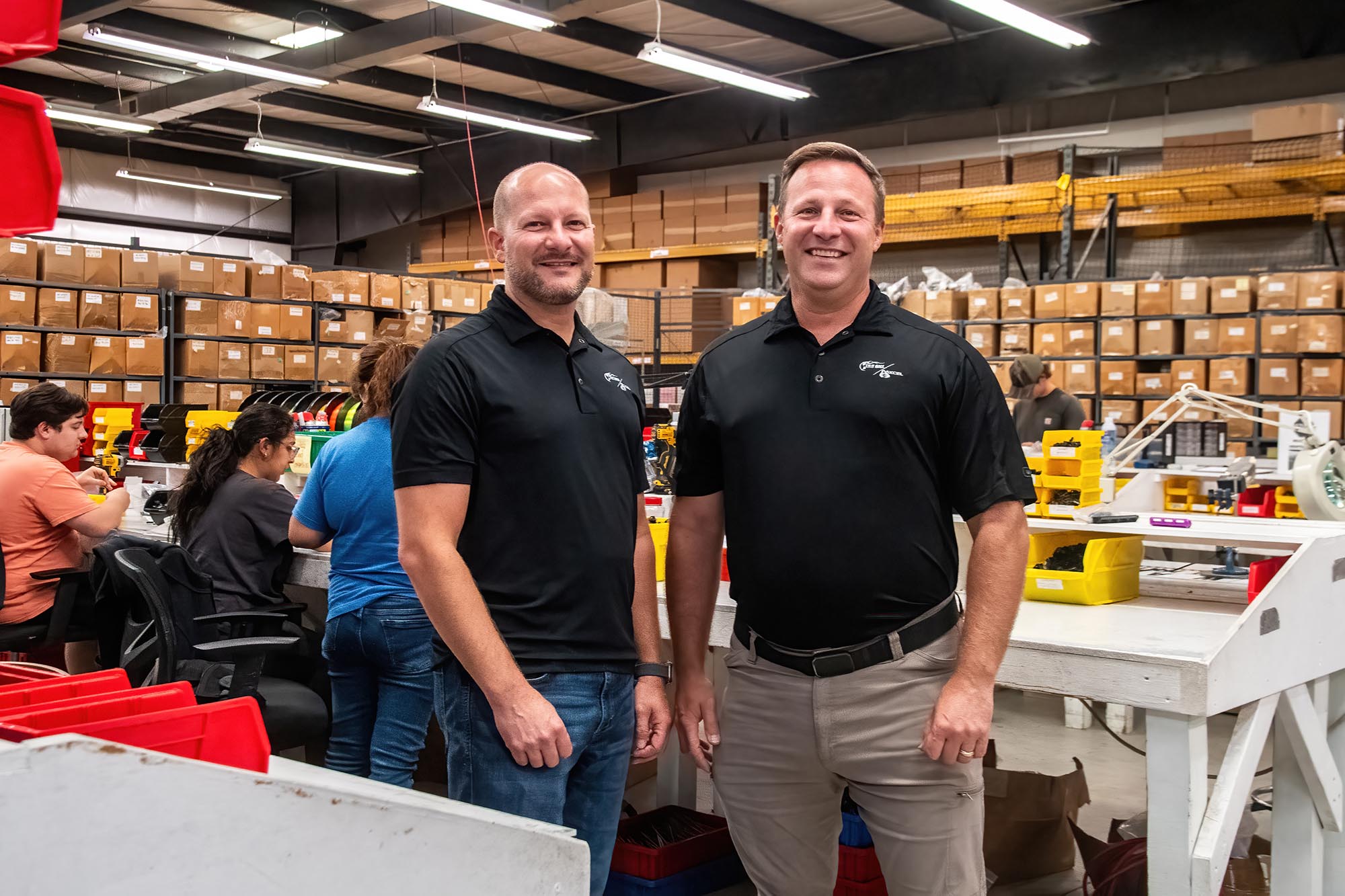 Ben and Brian Summers stand in the T.R.U. Ball/AXCEL Archery shop with employees working in the background. 