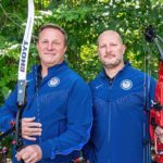 Ben Summers stands holding an Olympic style recurve bow with his brother Brian Summers holding a compound bow.