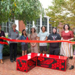 A large group of people hold up a red ribbon in preparation for a ribbon cutting.